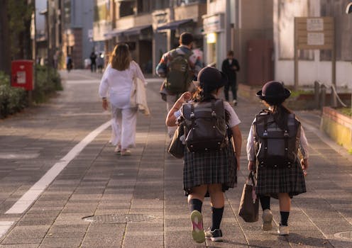 Two schoolgirls in uniform walk down a street with backpacks in an urban setting at sunset.